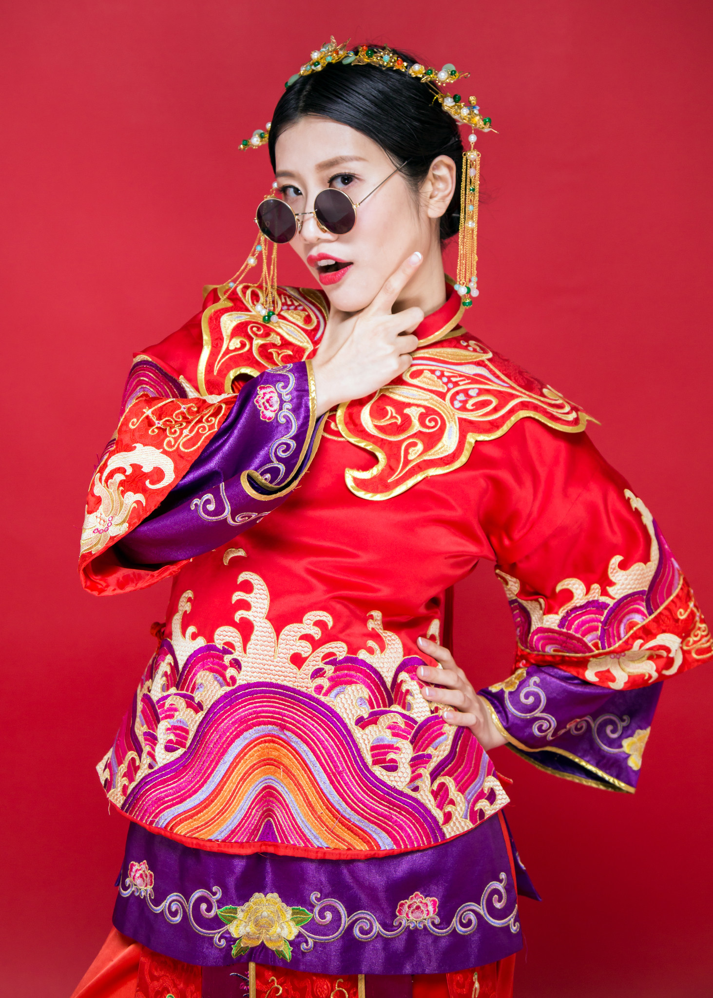 Original portrait with round sunglasses, ornate red, gold and purple clothing, and a playful hand-at-cheek pose against a red background.